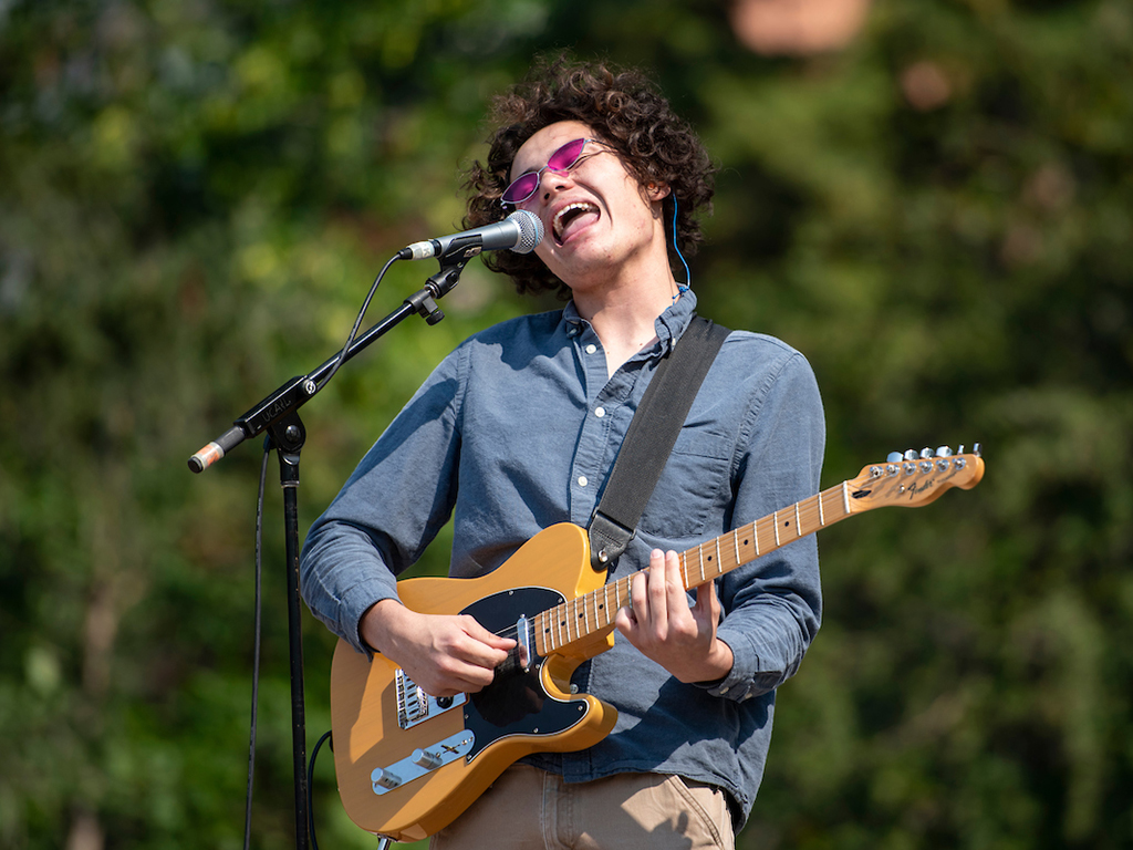 A student sings into a microphone while playing guitar on the Oval. 