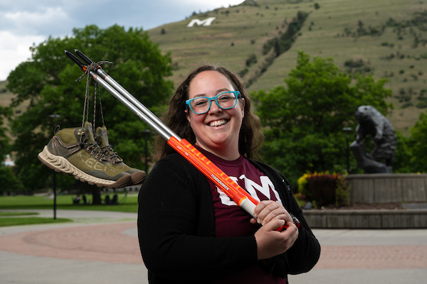 Brittany smiles for a portrait holding hiking poles with a pair of hiking boots dangling from their shoelaces from the hiking poles along the Oval on the University of Montana campus in Missoula, MT.