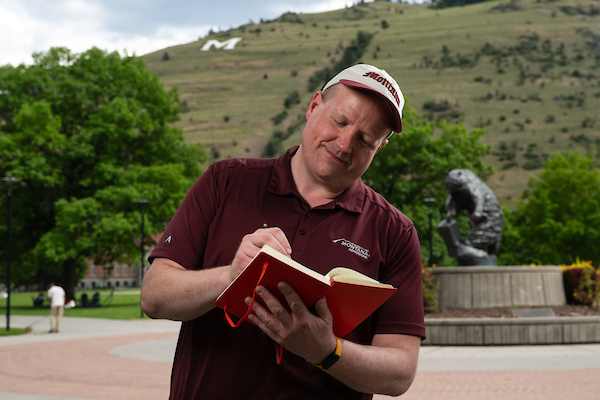 Colin, wearing University of Montana apparel, stands and writes in a notebook in front of the grizzly bear statue along the Oval on the University of Montana campus in Missoula, MT.