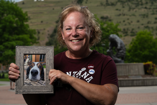 Kate smiles for a portrait wearing UM apparel in front of the grizzly bear statue along the Oval on the University of Montana campus in Missoula, MT and is holding a framed portrait of a dog.