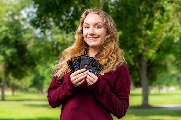 Tara stands for a portrait wearing University of Montana apparel, holding 4 "Magic, the Gathering" cards in her hands along the Oval on the UM campus in Missoula, MT.