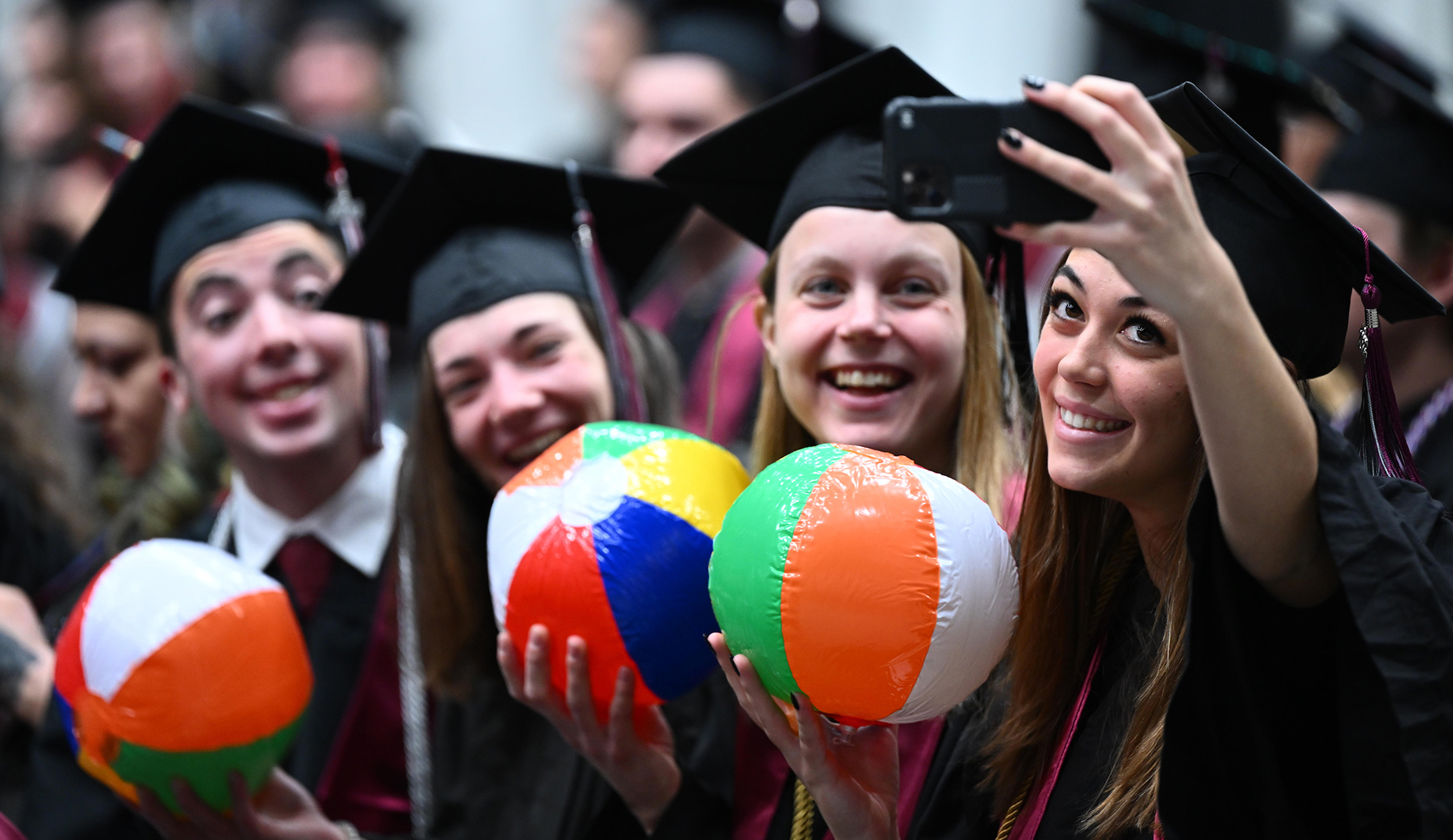 Students at Commencement smile while holding beach balls as the student on the right snaps a selfie of the group