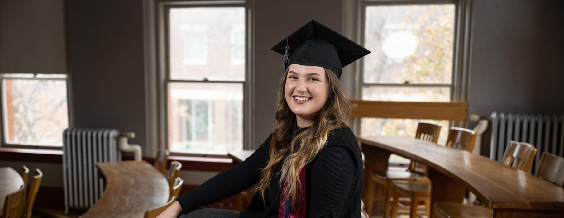 UM graduate Shanna Madison sits on a desk in Rankin Hall wearing her mortar board cap and smiles at the camera